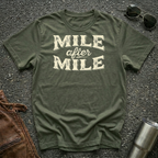 A military green graphic t-shirt laid flat on asphalt with sunglasses, a brown leather jacket, and a metal tumbler nearby. The shirt features the text 'MILE after MILE' in distressed cream lettering.