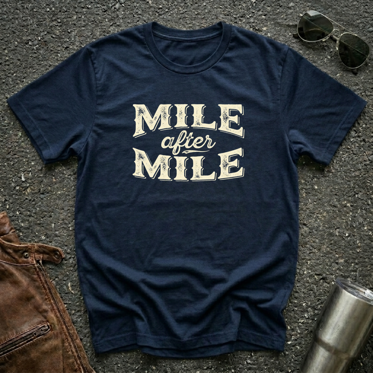 A navy graphic t-shirt laid flat on asphalt with sunglasses, a brown leather jacket, and a metal tumbler nearby. The shirt features the text 'MILE after MILE' in distressed cream lettering.