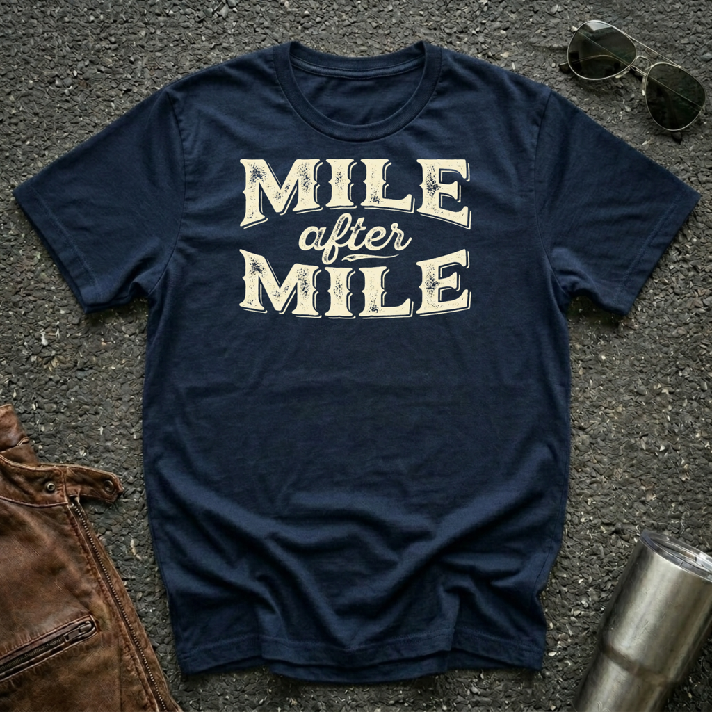 A navy graphic t-shirt laid flat on asphalt with sunglasses, a brown leather jacket, and a metal tumbler nearby. The shirt features the text 'MILE after MILE' in distressed cream lettering.