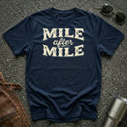 A navy graphic t-shirt laid flat on asphalt with sunglasses, a brown leather jacket, and a metal tumbler nearby. The shirt features the text 'MILE after MILE' in distressed cream lettering.