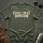 An military green t-shirt with the phrase 'CHASE THE NEXT HORIZON' printed in a beige serif font across the chest, laid flat on a dark asphalt surface with sunglasses, a brown leather bag, and a metal tumbler as styling props.