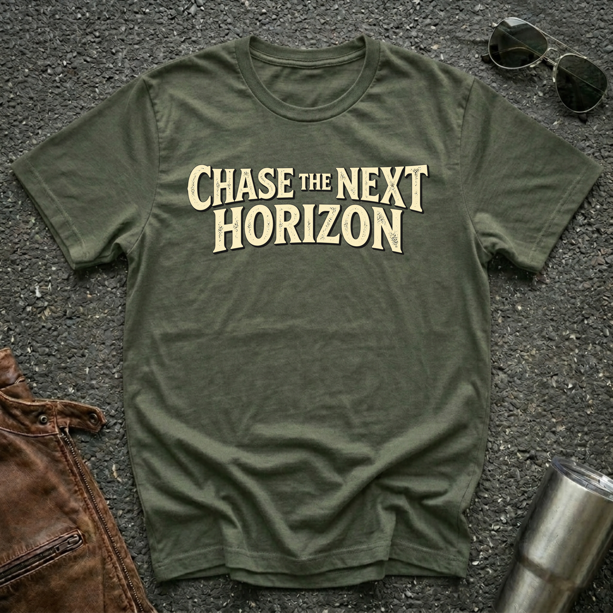 An military green t-shirt with the phrase 'CHASE THE NEXT HORIZON' printed in a beige serif font across the chest, laid flat on a dark asphalt surface with sunglasses, a brown leather bag, and a metal tumbler as styling props.
