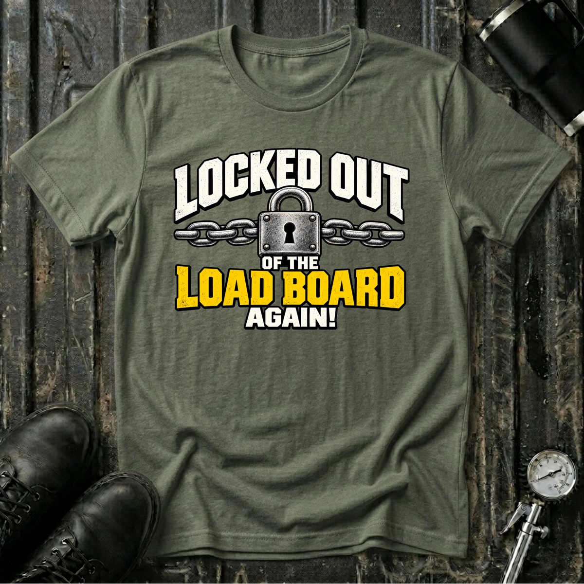 An military green graphic t-shirt laid flat on a dark rustic wooden background, styled with black leather boots and a bicycle pump. The t-shirt features a graphic print that reads 'LOCKED OUT OF THE LOAD BOARD AGAIN!' in white and yellow lettering with a padlock and chain illustration.