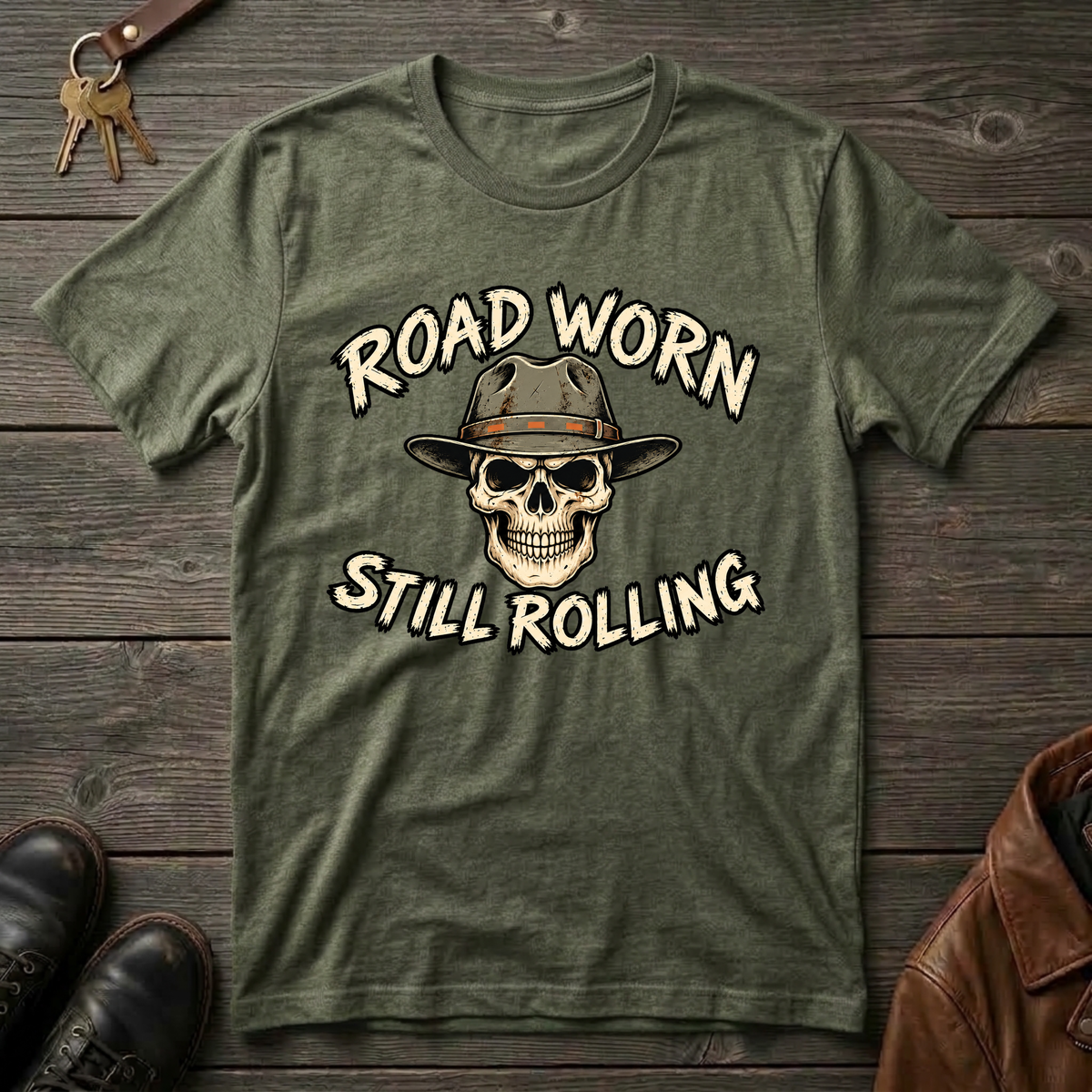 An military green graphic t-shirt laid flat on a dark wood background, featuring a skull wearing a cowboy hat with the text 'ROAD WORN STILL ROLLING' in cream lettering. Styled flat-lay props include keys at the top left, black shoes at the bottom left, and a brown leather jacket at the bottom right.