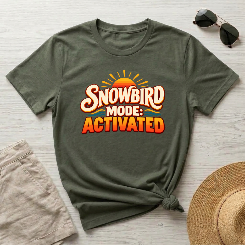 An olive green graphic t-shirt with a 'SNOWBIRD MODE: ACTIVATED' design, styled in a flat-lay arrangement on a light wood background with sunglasses, a straw hat, and beige shorts.