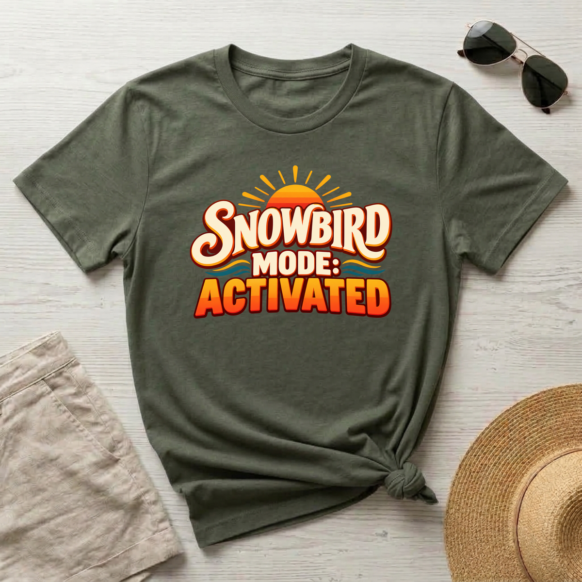 An olive green graphic t-shirt with a 'SNOWBIRD MODE: ACTIVATED' design, styled in a flat-lay arrangement on a light wood background with sunglasses, a straw hat, and beige shorts.
