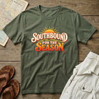 An military green short-sleeve t-shirt with a retro-style 'Southbound for the Season' graphic, styled in a flat-lay arrangement on a rustic wooden background alongside a map, brown sandals, and a light-colored shirt.