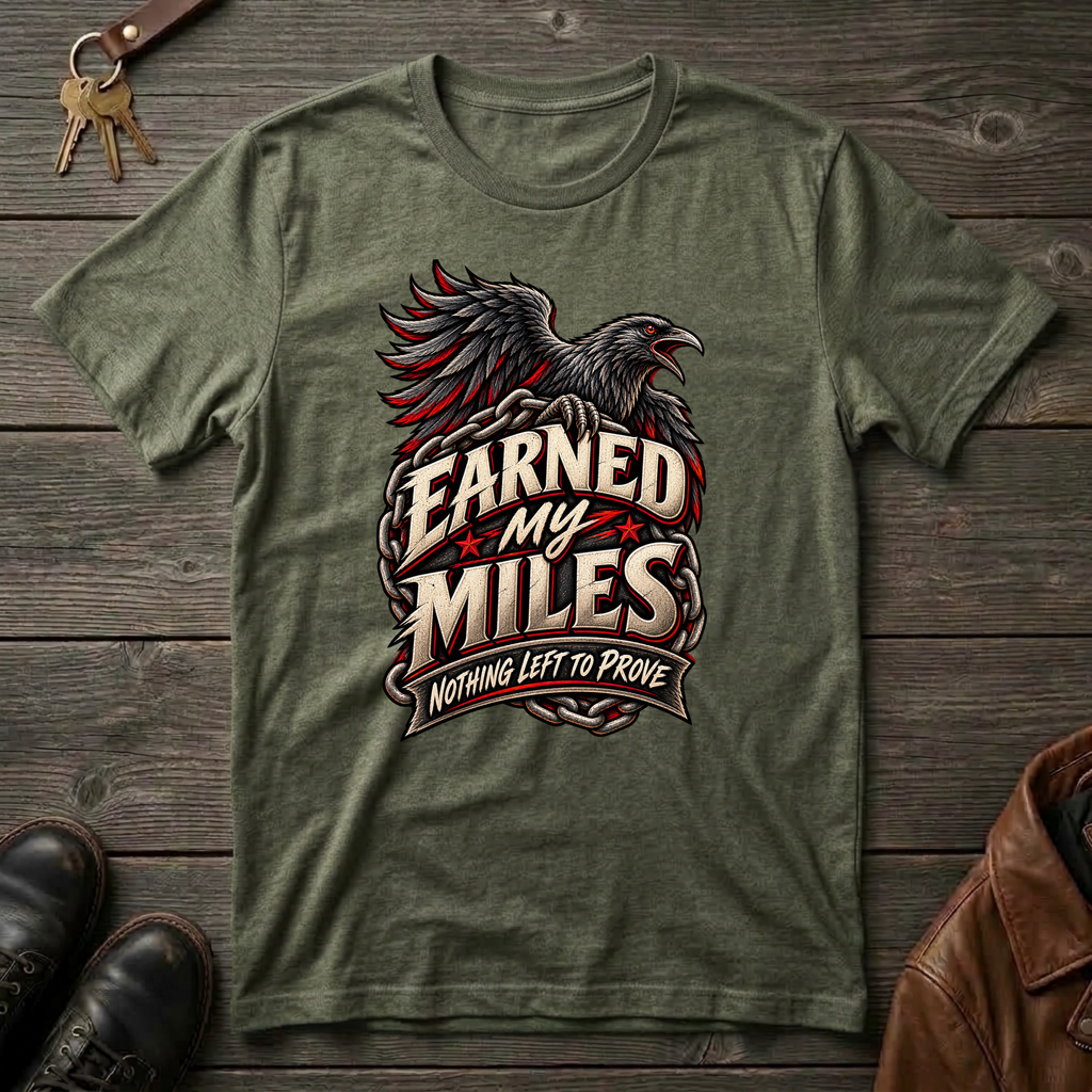 An military green graphic t-shirt featuring a raven and the text 'Earned My Miles Nothing Left to Prove', laid flat on a dark wood background with shoes and a leather jacket partially visible.