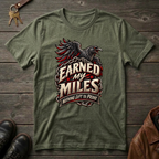 An military green graphic t-shirt featuring a raven and the text 'Earned My Miles Nothing Left to Prove', laid flat on a dark wood background with shoes and a leather jacket partially visible.