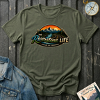An military green short-sleeve t-shirt with a retro mountain landscape graphic and the text 'The Riverstone Life Fueled by Freedom'. The shirt is styled in a flat-lay on a dark textured background with a denim jacket corner, a compass, and a 'Rocky Mountain National Park' stamp.