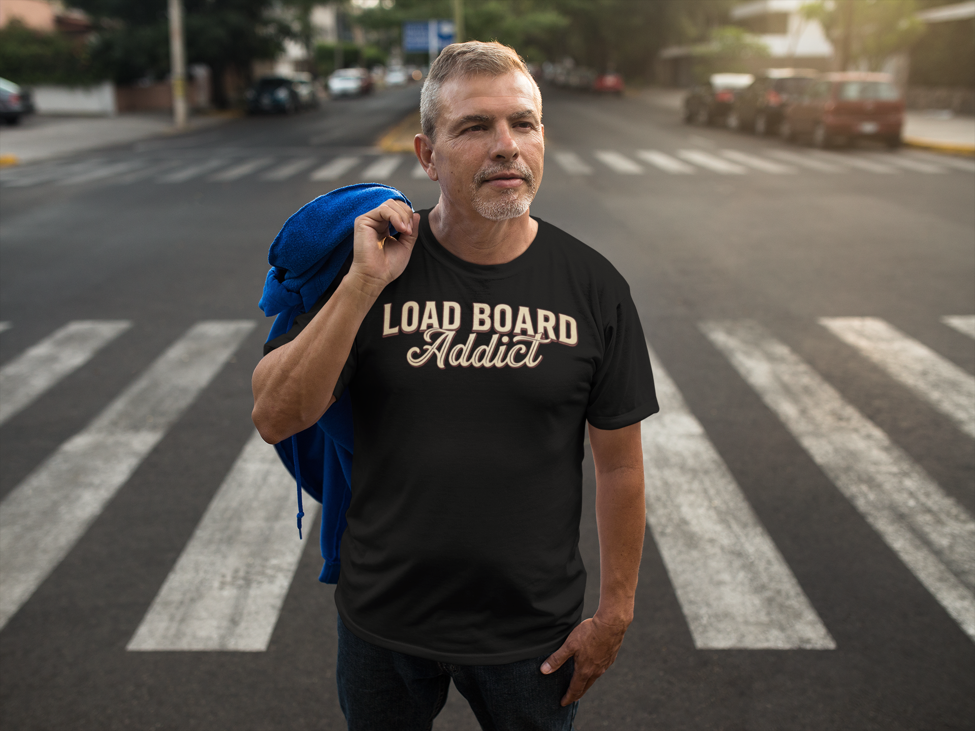 A man standing on a city crosswalk wearing a black t-shirt with 'LOAD BOARD Addict' printed in beige across the chest, holding a blue jacket over his shoulder.