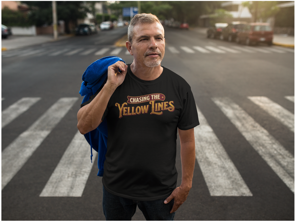 A man standing on a city crosswalk wearing a black graphic T-shirt that reads 'Chasing the Yellow Lines' in gold and red lettering, with a blue jacket draped over his shoulder. Cars and buildings are visible in the blurred background.