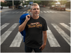 A man standing on a city crosswalk wearing a black graphic T-shirt that reads 'Chasing the Yellow Lines' in gold and red lettering, with a blue jacket draped over his shoulder. Cars and buildings are visible in the blurred background.