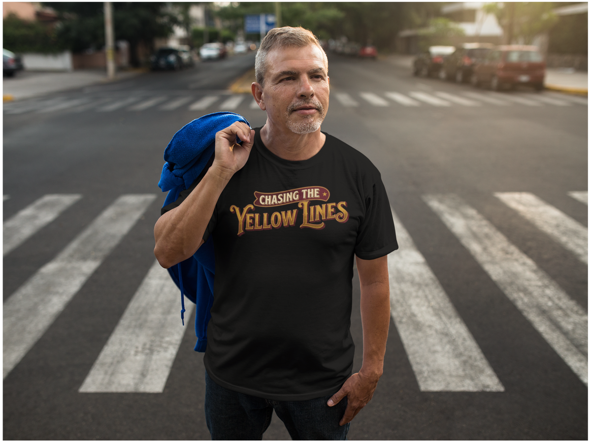 A man standing on a city crosswalk wearing a black graphic T-shirt that reads 'Chasing the Yellow Lines' in gold and red lettering, with a blue jacket draped over his shoulder. Cars and buildings are visible in the blurred background.