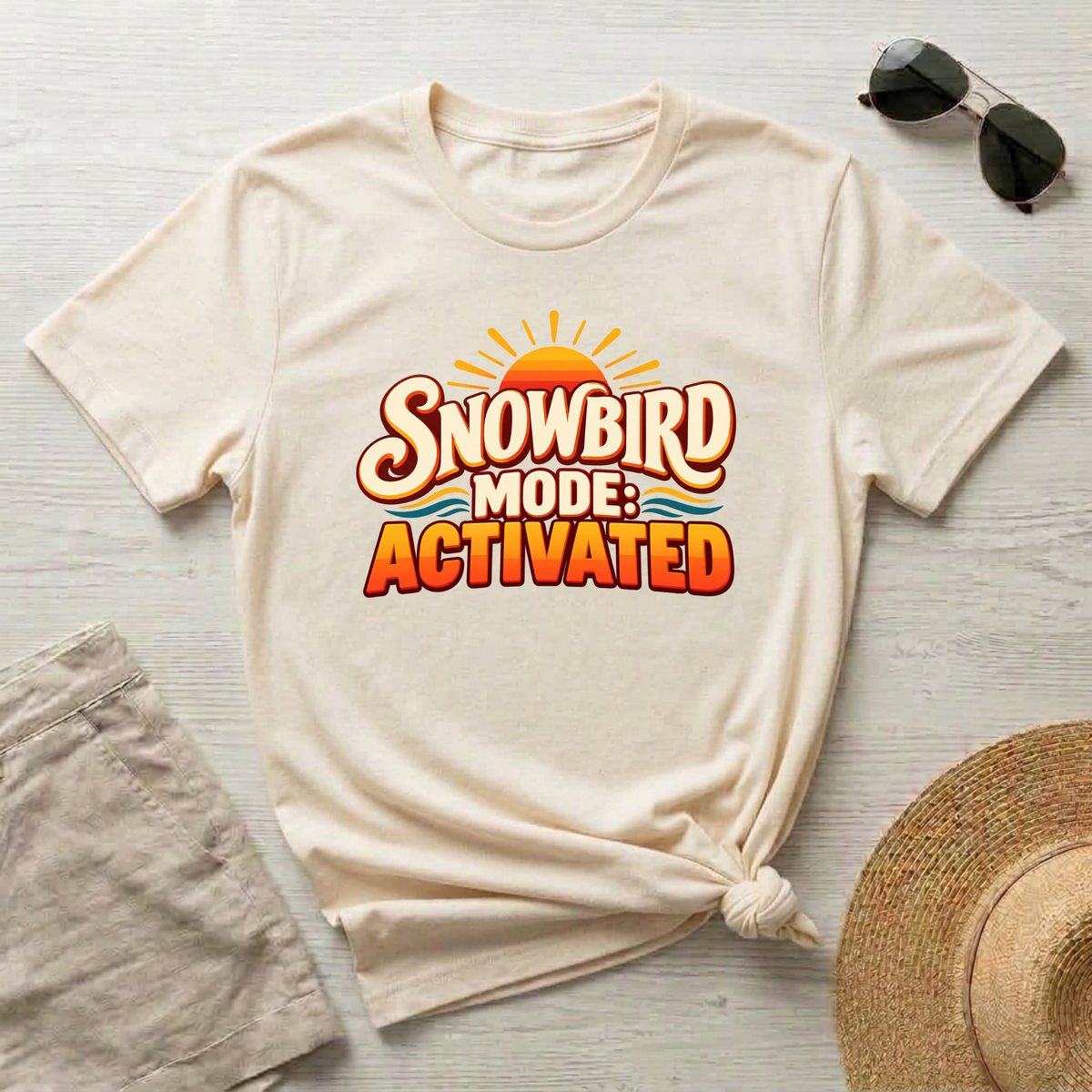 A natural-colored t-shirt with a retro-style graphic reading 'SNOWBIRD MODE: ACTIVATED' in orange and yellow, styled in a flat-lay with sunglasses, a straw hat, and linen shorts on a light wood background.