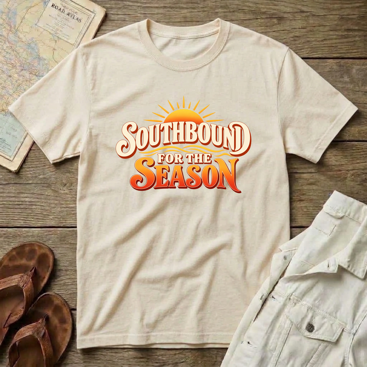 A natural color short-sleeve t-shirt laid flat on a rustic wooden surface, featuring a colorful graphic that reads 'SOUTHBOUND FOR THE SEASON' with a sunburst behind the text. The shirt is styled with folded white shorts, brown sandals, and a vintage map.