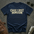 A navy blue crewneck t-shirt with the phrase 'CHASE THE NEXT HORIZON' printed in a beige serif font across the chest. The shirt is laid flat on a dark asphalt surface, styled with sunglasses, a brown leather jacket, and a metal tumbler.