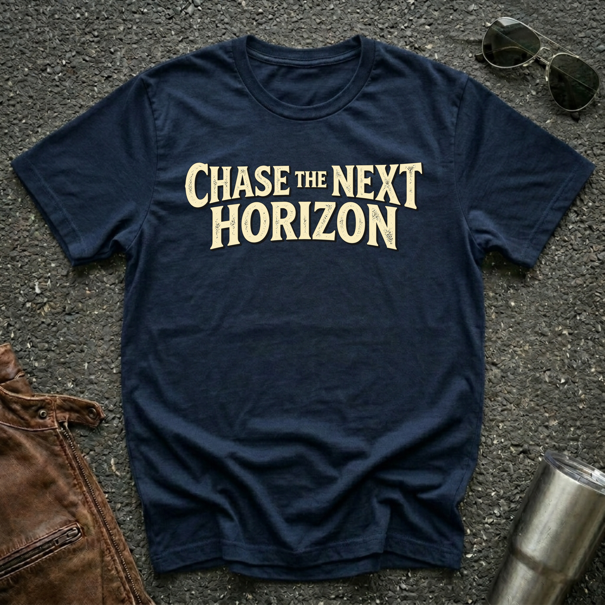 A navy blue crewneck t-shirt with the phrase 'CHASE THE NEXT HORIZON' printed in a beige serif font across the chest. The shirt is laid flat on a dark asphalt surface, styled with sunglasses, a brown leather jacket, and a metal tumbler.