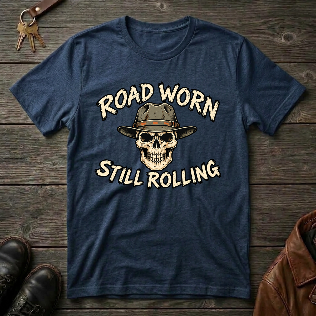 A navy blue graphic t-shirt laid flat on a rustic dark wood background, featuring a skull wearing a cowboy hat with the text 'ROAD WORN STILL ROLLING' in cream. Decorative props including keys, brown leather boots, and a brown leather jacket are partially visible around the shirt.