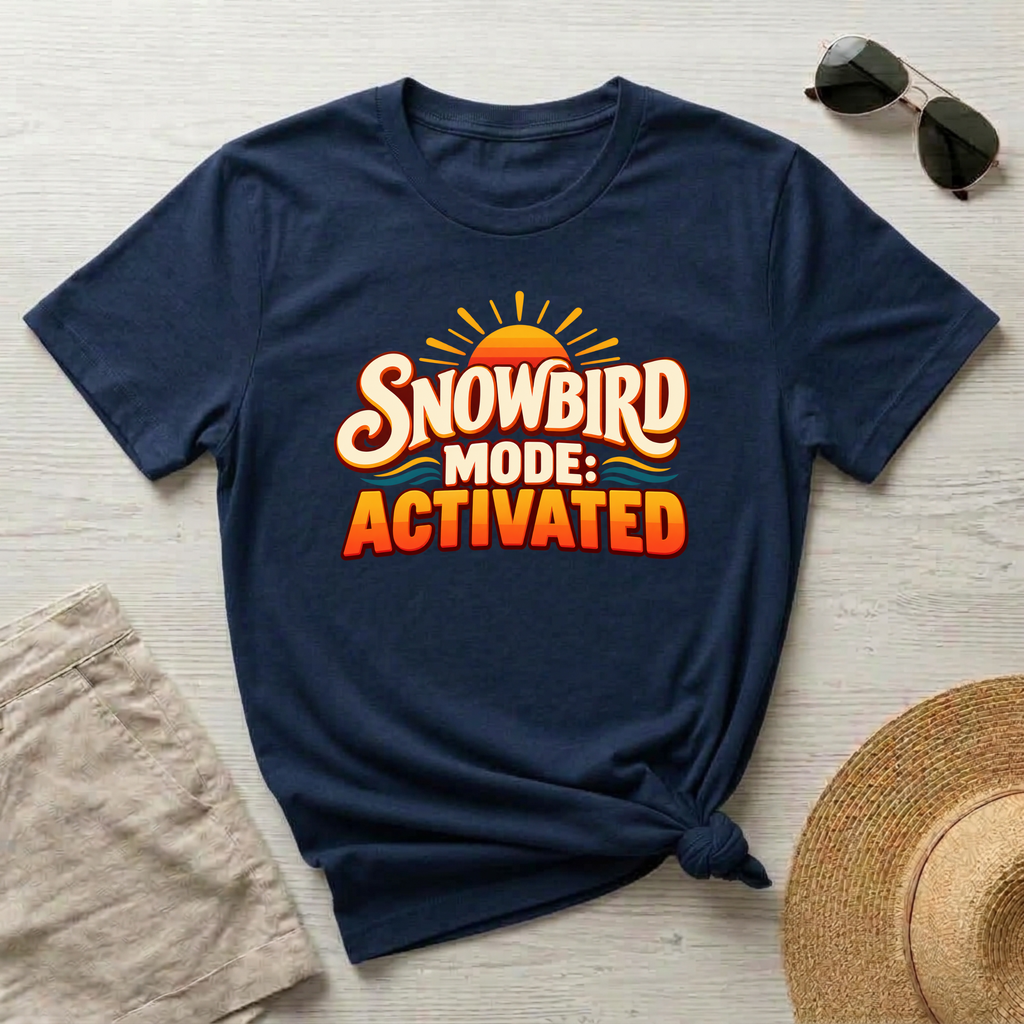 A navy blue short-sleeve t-shirt with a 'SNOWBIRD MODE: ACTIVATED' graphic print, styled in a flat-lay arrangement with sunglasses, beige shorts, and a straw hat on a light wood background.