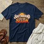 A navy blue short-sleeve t-shirt with a retro-style graphic reading 'SOUTHBOUND FOR THE SEASON' in white and orange lettering with a yellow sunburst, styled in a flat lay on a wooden surface with sandals, a map, and light-colored shorts nearby.