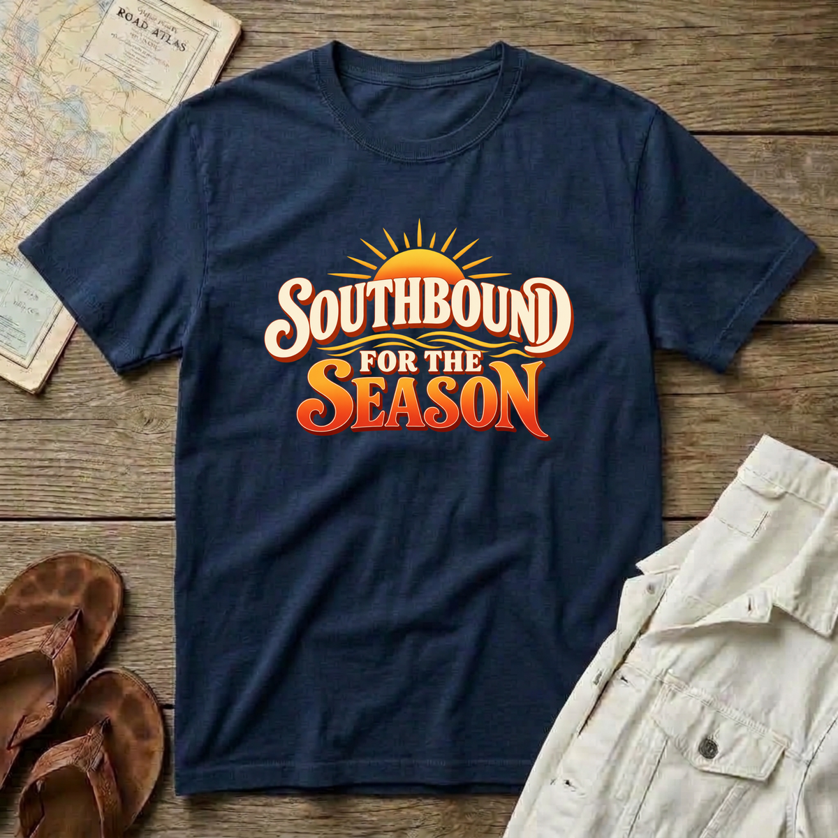 A navy blue short-sleeve t-shirt with a retro-style graphic reading 'SOUTHBOUND FOR THE SEASON' in white and orange lettering with a yellow sunburst, styled in a flat lay on a wooden surface with sandals, a map, and light-colored shorts nearby.