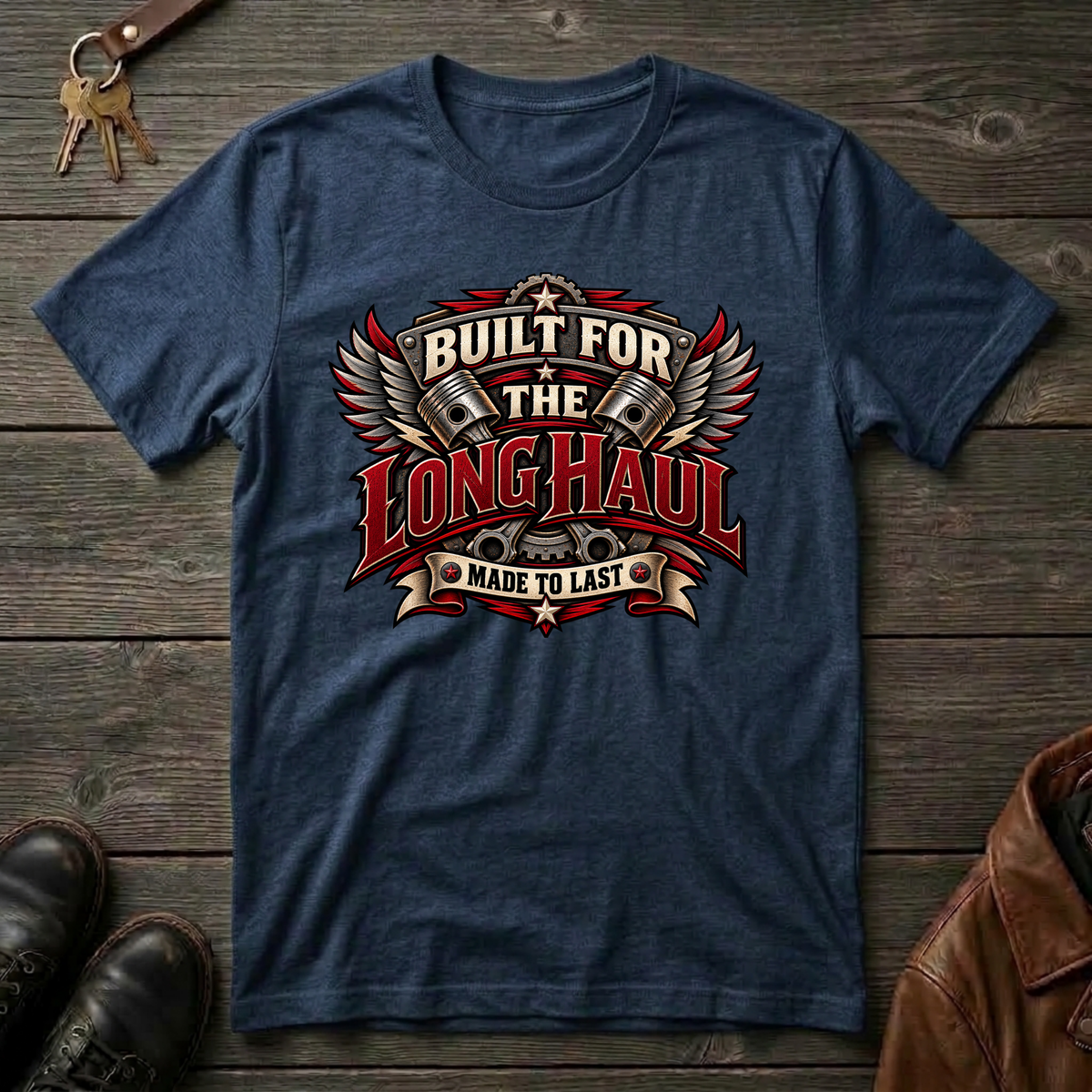 A navy graphic t-shirt laid flat on a rustic dark wood background, styled with keys, brown leather shoes, and a brown leather jacket. The t-shirt features a large graphic on the chest with wings, pistons, and the text 'BUILT FOR THE LONG HAUL MADE TO LAST'.