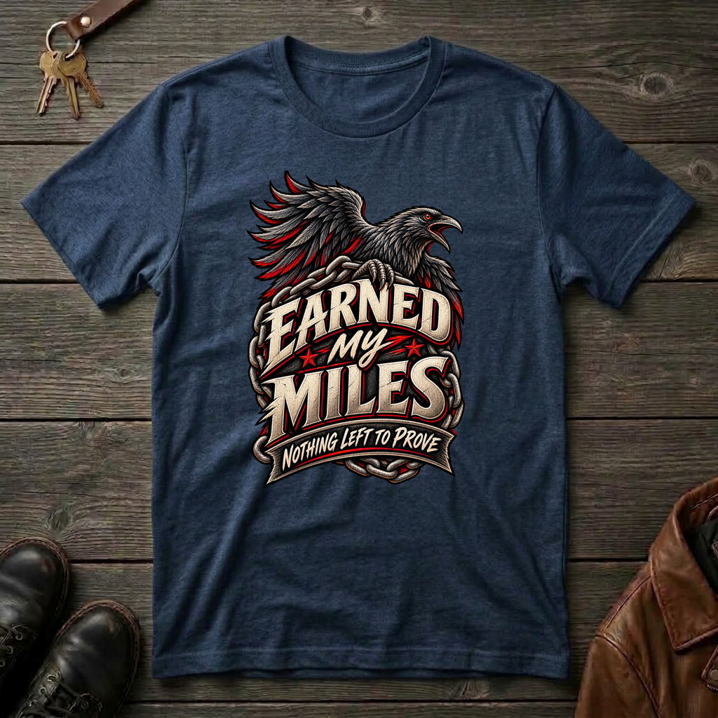 A navy blue graphic t-shirt featuring a raven and the text 'Earned My Miles Nothing Left to Prove', laid flat on a dark wood background with boots, keys, and a leather jacket nearby.