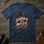 A navy blue graphic t-shirt featuring a raven and the text 'Earned My Miles Nothing Left to Prove', laid flat on a dark wood background with boots, keys, and a leather jacket nearby.
