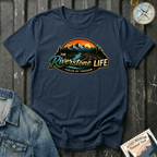 A navy graphic t-shirt laid flat on a dark textured background, featuring a scenic mountain and river design with the text 'The Riverstone Life' and 'Fueled by Freedom'. A compass and a Rocky Mountain National Park stamp are styled as props around the shirt.