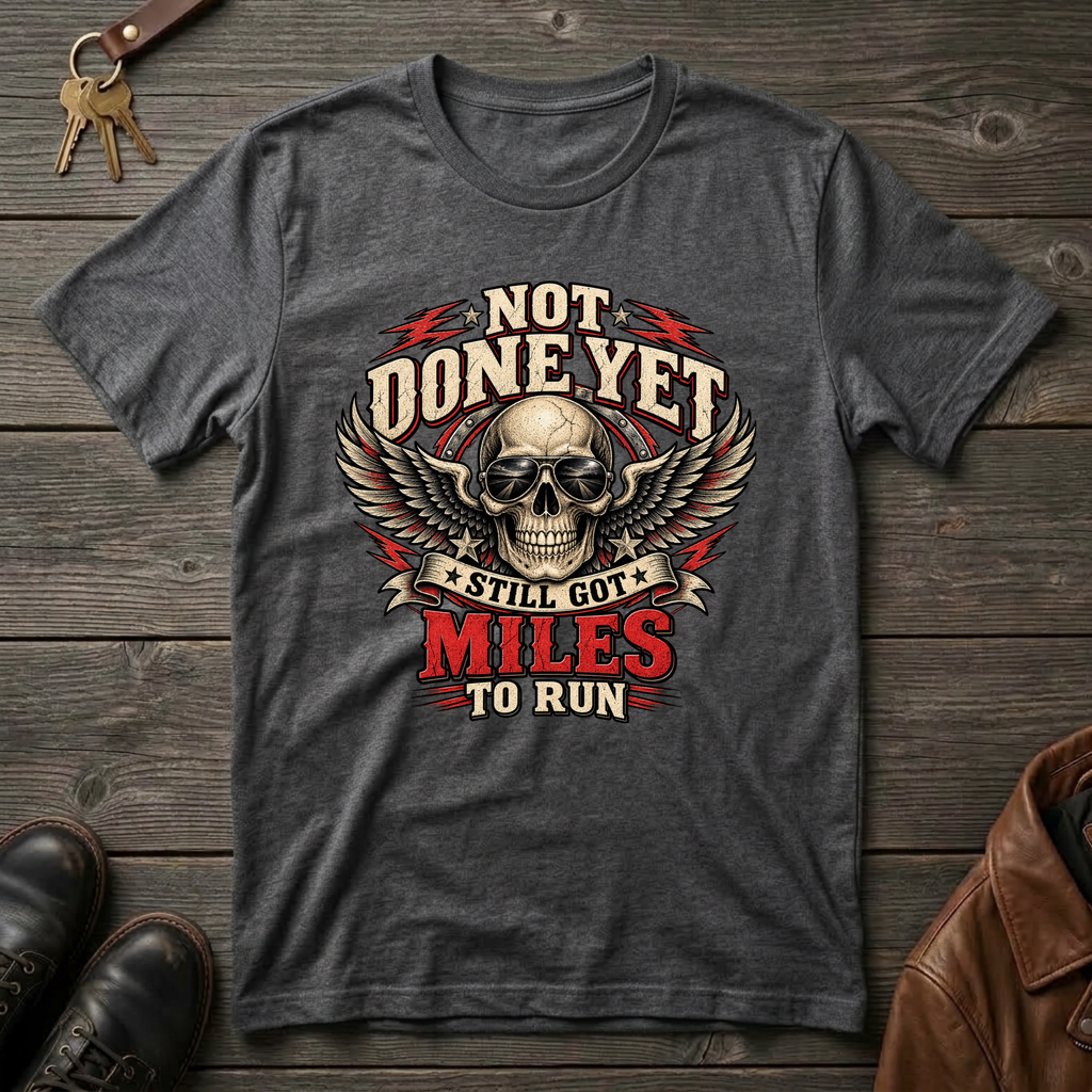 A dark heather heather graphic t-shirt laid flat on a rustic wooden background, featuring a skull with wings and the text 'NOT DONE YET STILL GOT MILES TO RUN'.