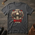 A dark heather heather graphic t-shirt laid flat on a rustic wooden background, featuring a skull with wings and the text 'NOT DONE YET STILL GOT MILES TO RUN'.