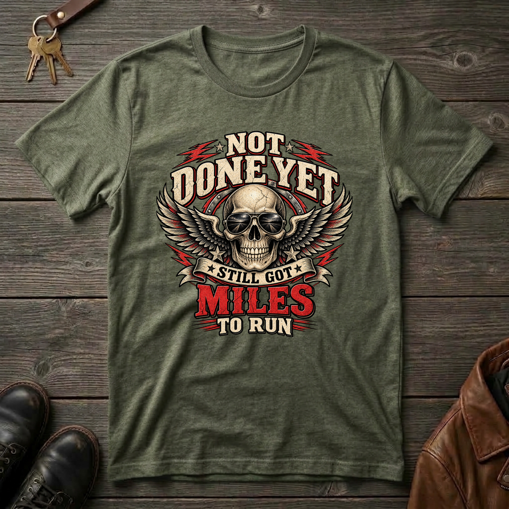 An military green graphic t-shirt laid flat on a dark wood background, styled with keys, leather shoes, and a brown leather jacket. The t-shirt features a graphic of a skull with wings and the text 'NOT DONE YET', 'STILL GOT', 'MILES', and 'TO RUN'.