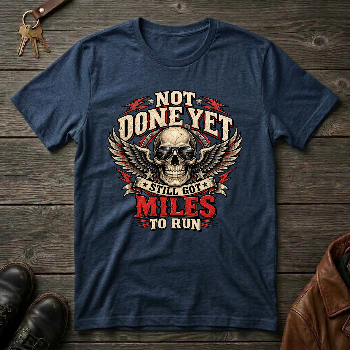 A navy graphic t-shirt laid flat on a rustic dark wood background, styled with keys, brown leather shoes, and a brown leather jacket. The t-shirt features a large graphic of a skull with wings and the text 'NOT DONE YET STILL GOT MILES TO RUN'.
