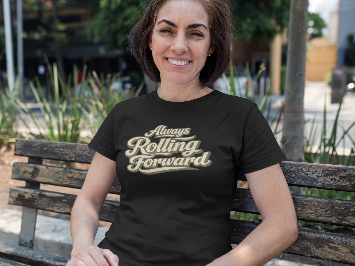 A woman sitting on a wooden park bench wearing a black t-shirt with a gold graphic that reads 'Always Rolling Forward'.