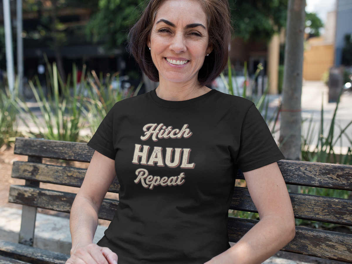 A woman sitting on a wooden park bench wearing a black t-shirt with the text 'Hitch HAUL Repeat' in a beige and brown distressed font. The background shows a blurred outdoor park setting with greenery and a street.