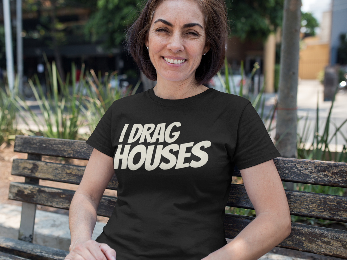 A woman sitting on a wooden bench outdoors wearing a black t-shirt with the text 'I DRAG HOUSES' printed in bold white letters.