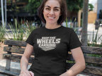 A woman sitting on a wooden park bench wearing a black t-shirt with a white graphic that reads 'KEEP THE WHEELS EARNING' 
