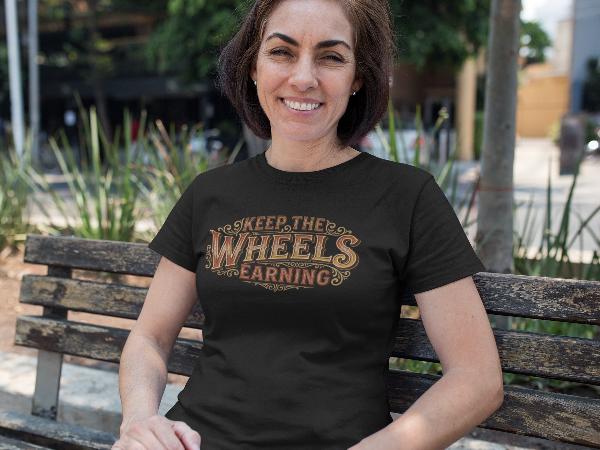 A woman sitting on a wooden bench outdoors wearing a black graphic t-shirt with the text 'KEEP, THE WHEELS EARNING' in a distressed brown and gold font.