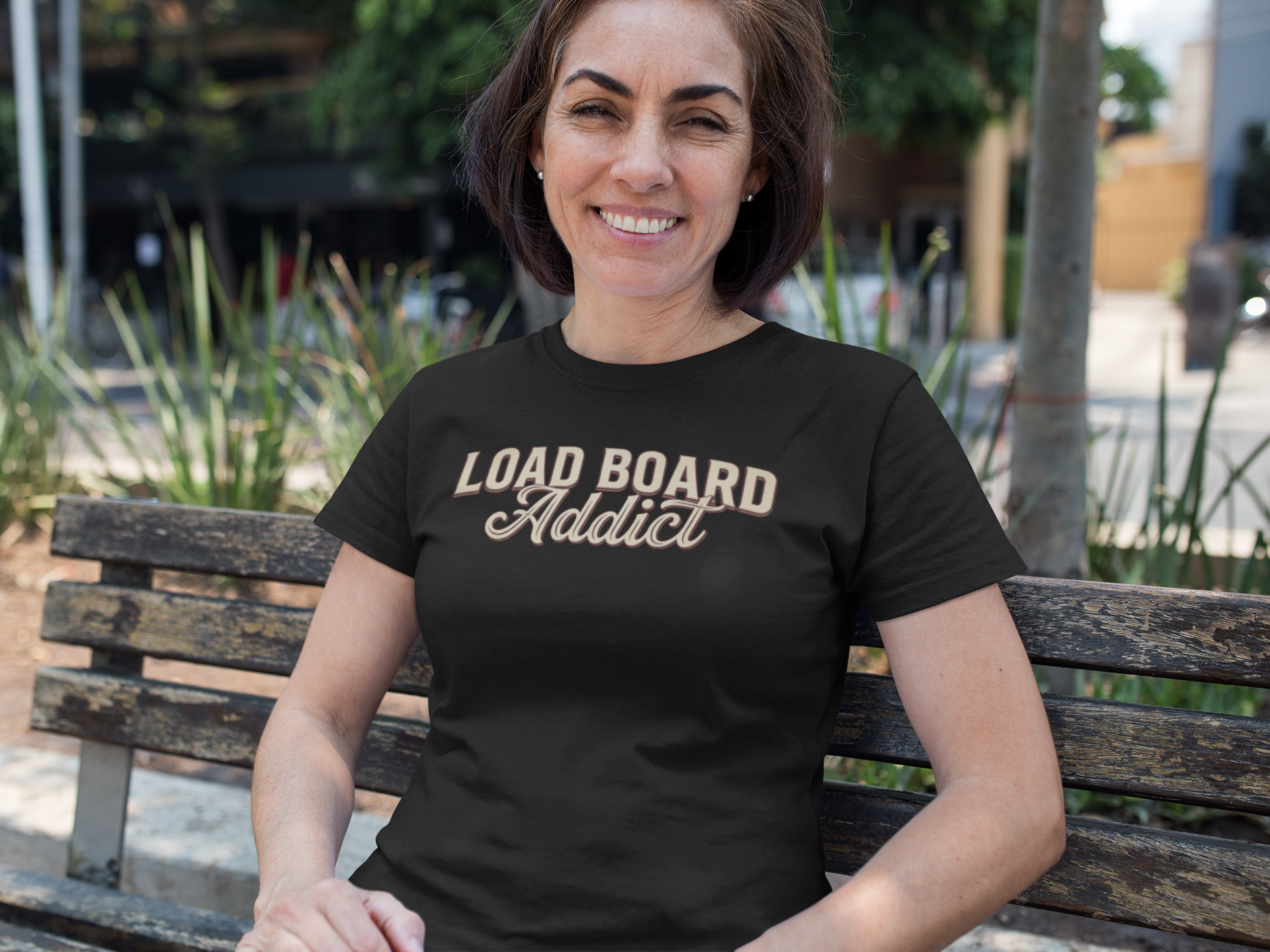 A woman sitting on a wooden bench outdoors wearing a black t-shirt with 'LOAD BOARD Addict' printed in beige across the chest.