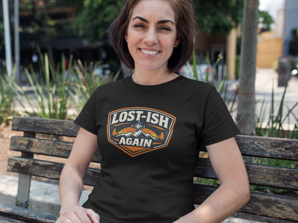 A woman sitting on a wooden bench outdoors wearing a black t-shirt with a graphic that reads 'LOST-ISH AGAIN' in white, orange, and brown, with a mountain and tree illustration.