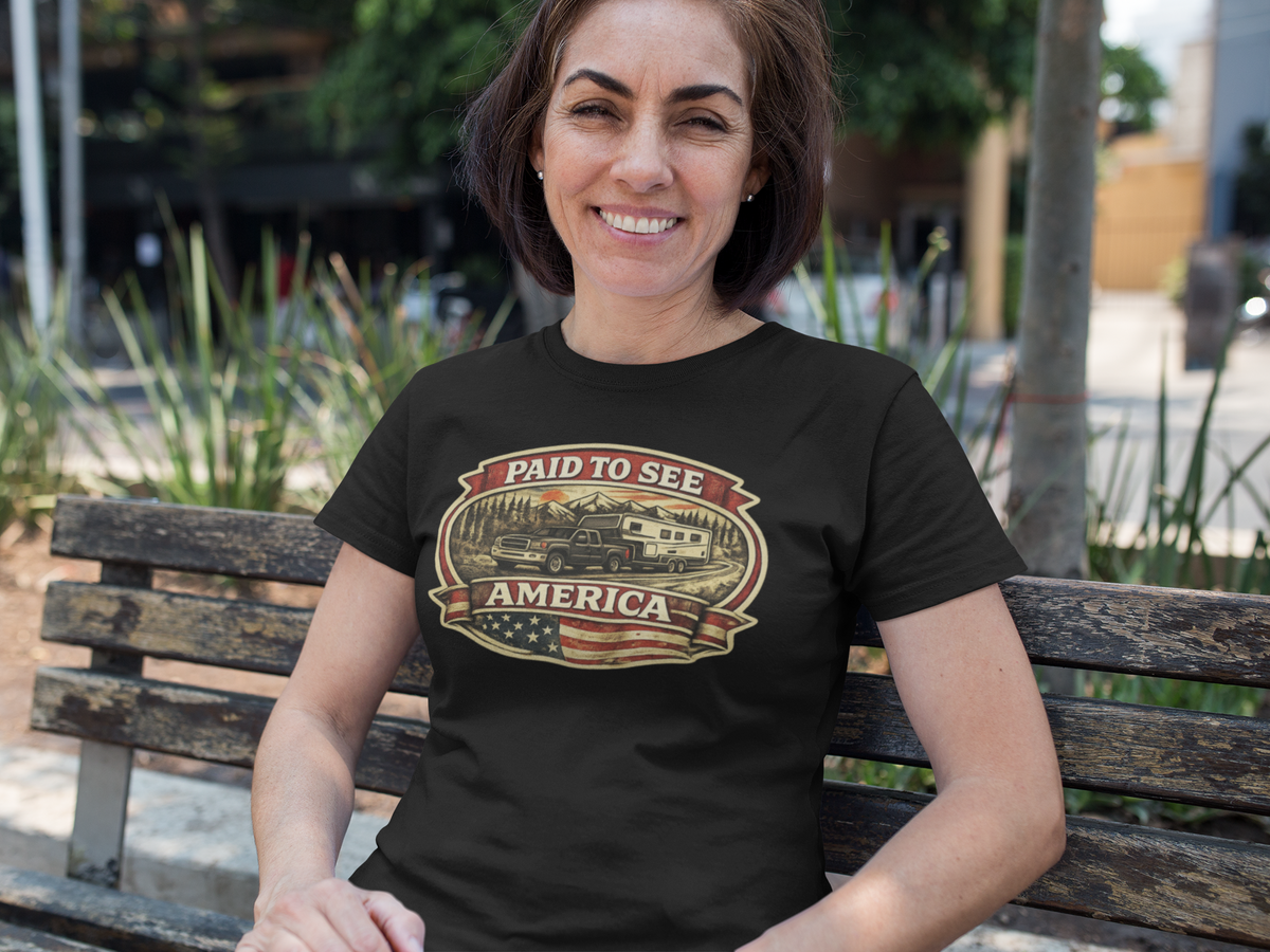 A woman sitting on a wooden bench outdoors wearing a black t-shirt with a vintage-style American flag and camper graphic that reads 'PAID TO SEE AMERICA'.