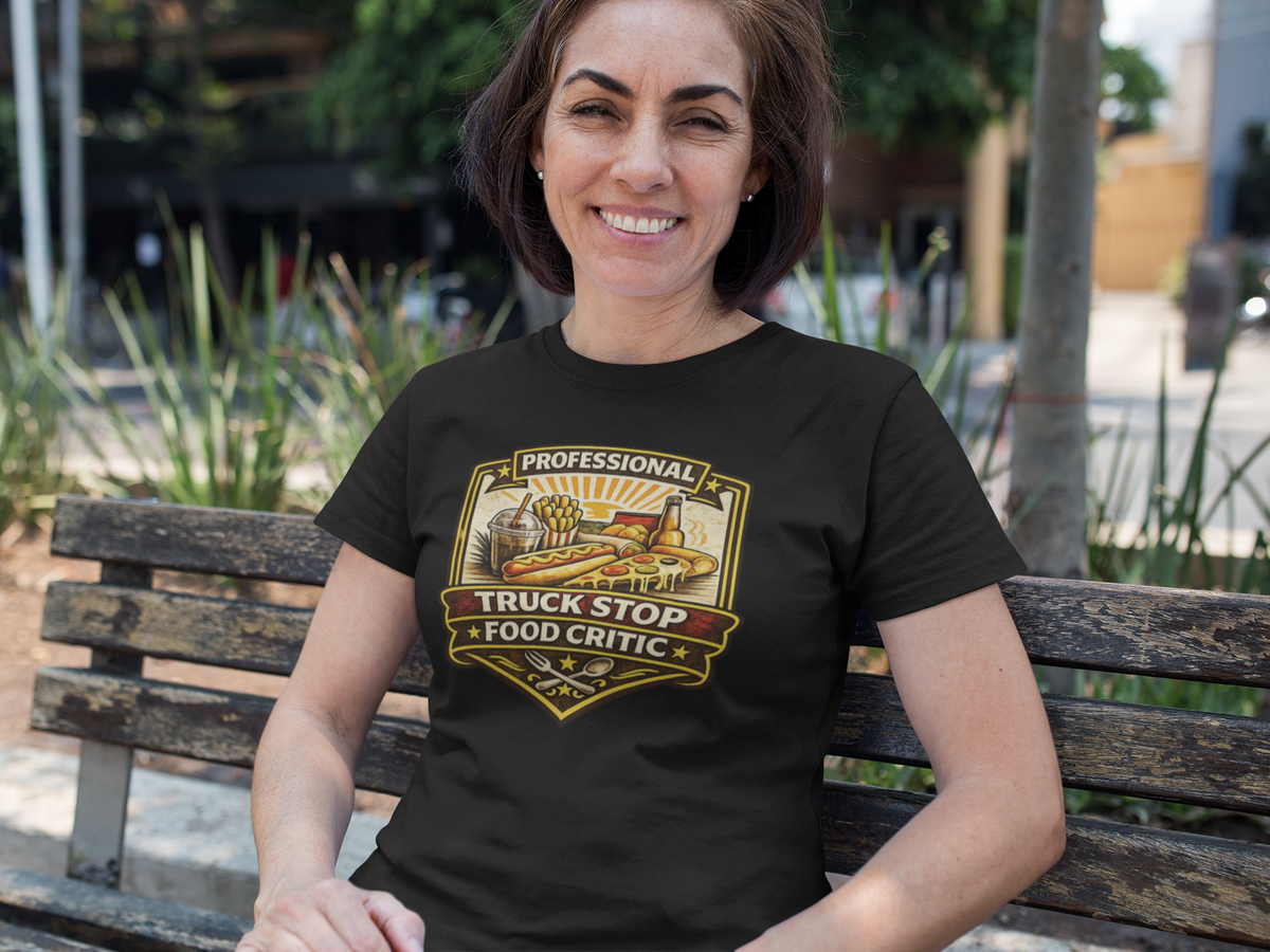 A smiling woman sits on a wooden park bench wearing a black graphic t-shirt with a 'PROFESSIONAL TRUCK STOP FOOD CRITIC' The background shows a blurred outdoor park setting with greenery and a street.