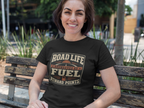 A woman sitting on a wooden park bench wearing a black t-shirt with a brown and gold graphic that reads 'Road Life Sponsored By Fuel Reward Points'