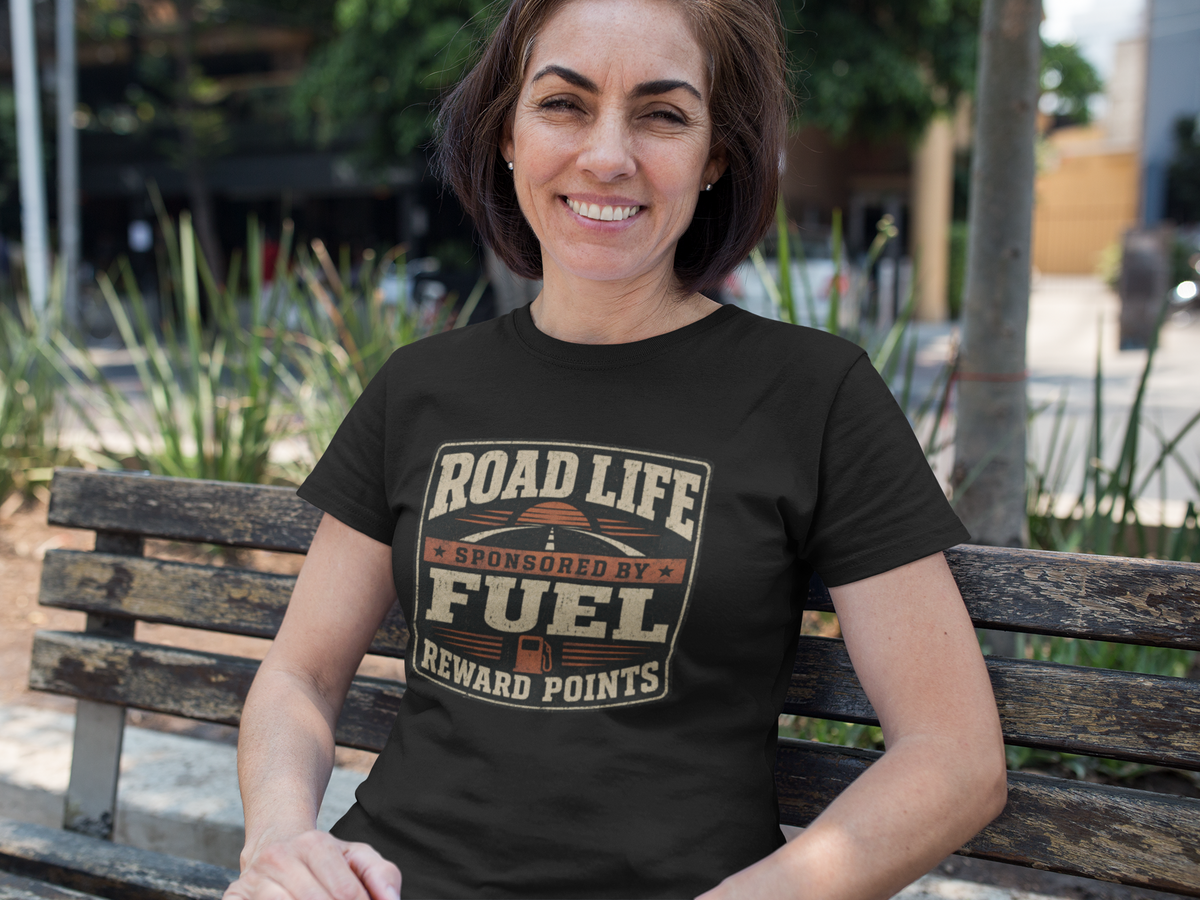 A woman sitting on a wooden park bench wearing a black t-shirt with a brown and gold graphic that reads 'Road Life Sponsored By Fuel Reward Points'