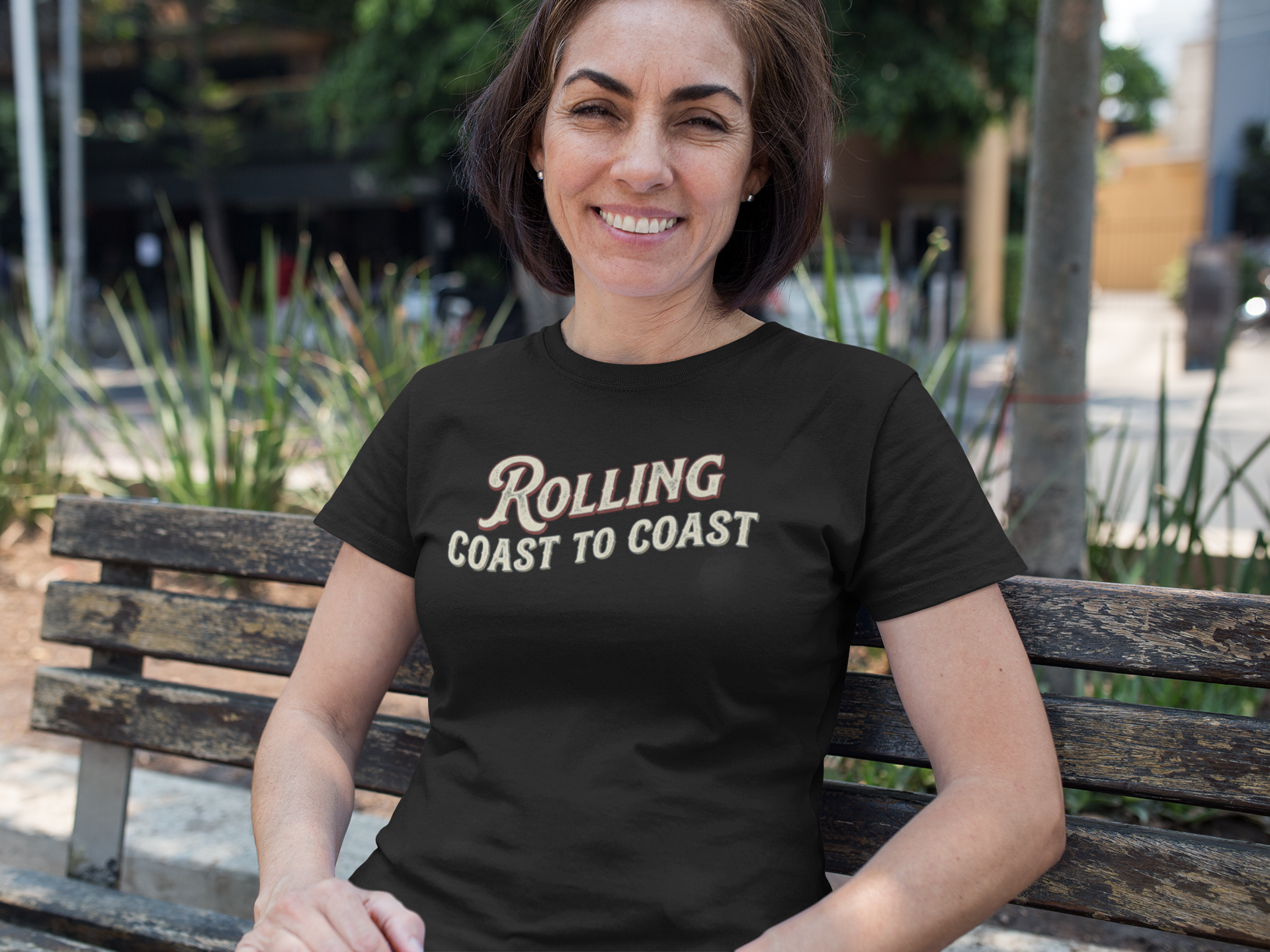 A woman sitting on a wooden bench outdoors wearing a black graphic t-shirt with the text 'Rolling Coast to Coast' in a distressed brown and gold font.