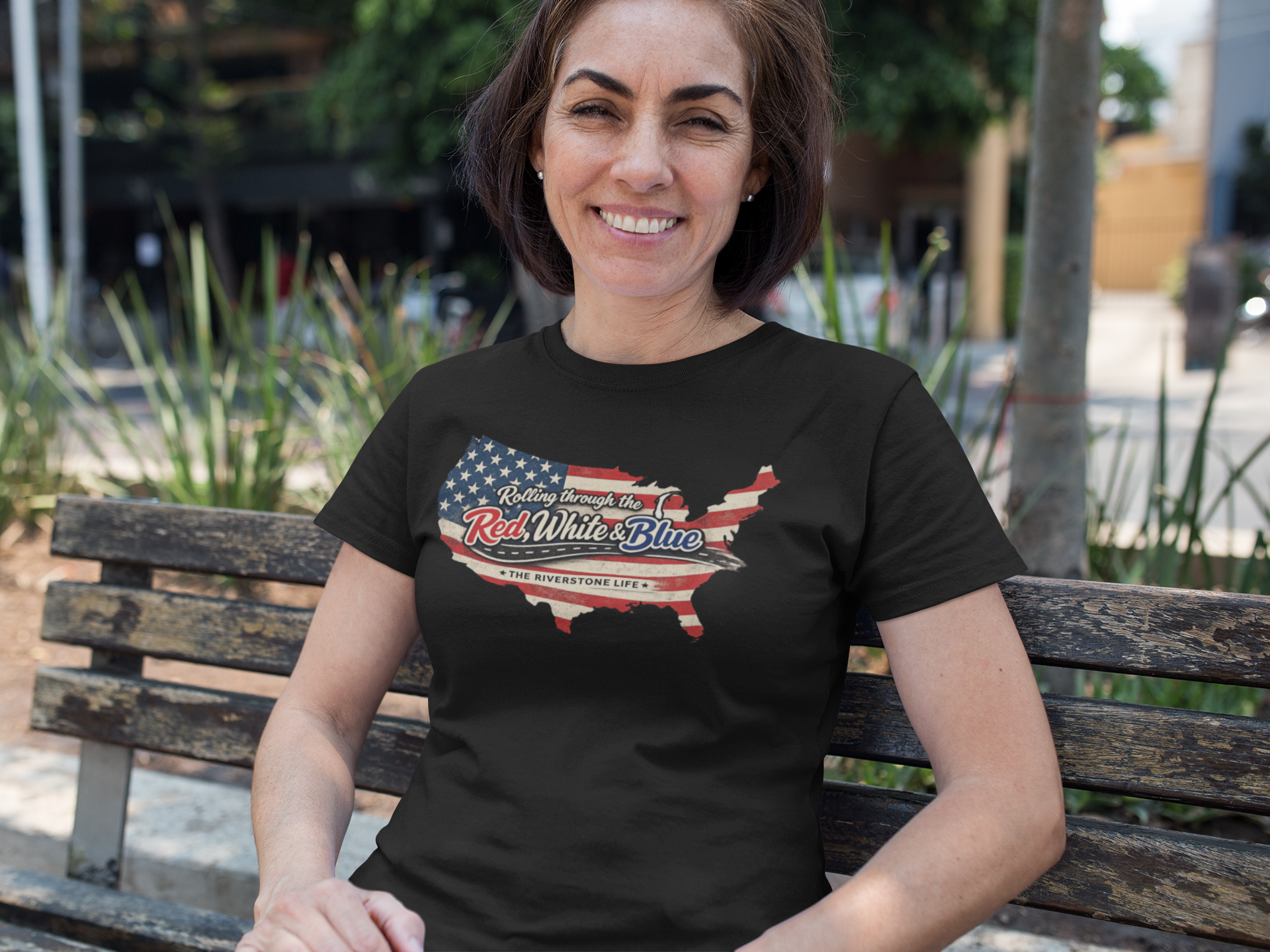 A woman sitting on a wooden bench outdoors wearing a black t-shirt with a graphic of the United States map filled with the American flag and the text 'Rolling through the Red, White & Blue' and 'THE RIVERSTONE LIFE'.