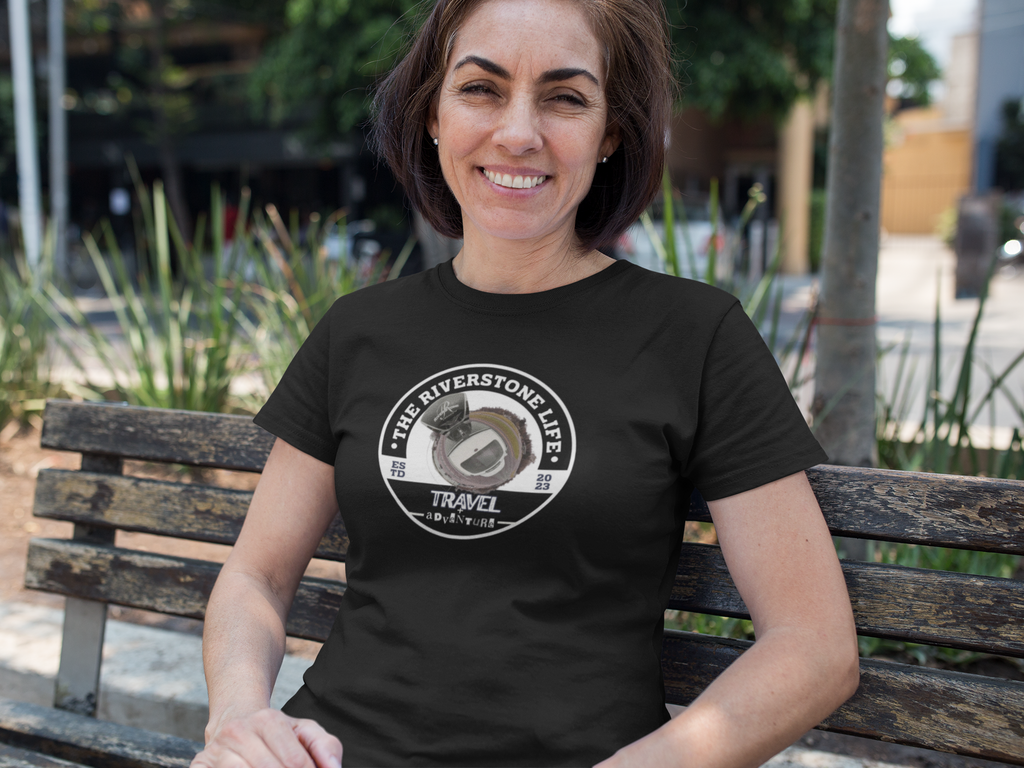 A woman sitting on a wooden bench outdoors wearing a black t-shirt with a circular white graphic that reads 'THE RIVERSTONE LIFE TRAVEL AND ADVENTURE' features a stylized map icon.