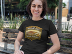 A smiling woman sits on a wooden park bench wearing a black graphic t-shirt with a 'The Roller Grill Never Judges' design. The background shows a blurred outdoor park setting with greenery background.