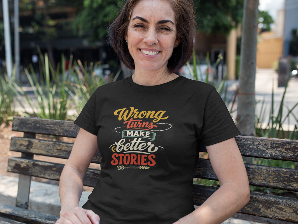 A woman sitting on a wooden bench outdoors wearing a black graphic t-shirt with the text 'Wrong Turns Make Better Stories' in yellow, white, and red lettering.